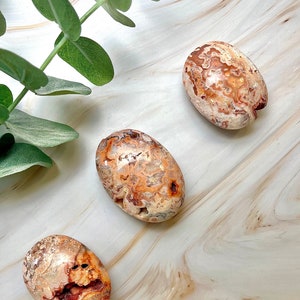 May include: Three smooth, oval-shaped agate stones with a white, orange, and brown color pattern. The stones are on a light brown surface.