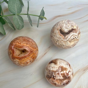 May include: Three agate spheres with a natural, brown, white, and pink color pattern. The spheres have a hollow center with a crystal formation.