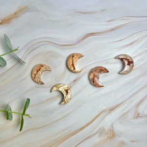 May include: Five crescent moon shaped polished stones with a light brown and gold color. The stones are arranged on a white marble surface.