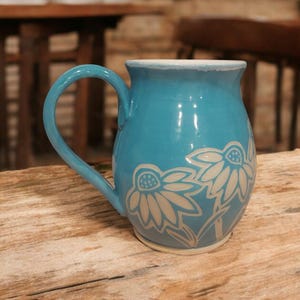 Puede incluir: Una taza de cerámica turquesa con un asa grande y un diseño floral blanco. La taza tiene un borde ligeramente texturizado y un acabado liso y brillante. La taza mide aproximadamente 10 cm de alto.