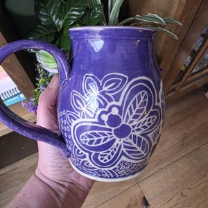 May include: A purple ceramic mug with a large handle and a white floral design. The mug is hand-painted with a glossy finish. The mug is held by a person.