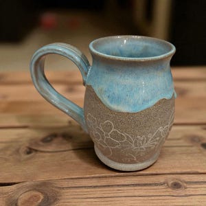 May include: Handmade ceramic mug with a light blue glazed top and a grey, textured base. The base features a white floral design. The mug has a curved handle and is set on a wooden surface.