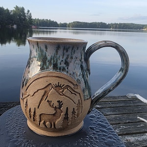 May include: A handcrafted ceramic mug with a nature-inspired design. The mug showcases a detailed relief of a deer, mountains, and trees. The upper part has a blue and green glaze, while the lower section is a natural tan colour. The mug is set against a lake backdrop.