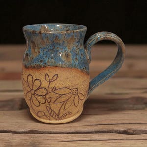 May include: Handmade ceramic mug with a blue and brown glazed top and a tan base. The base features a floral design. The mug has a curved handle and is suitable for hot or cold beverages. A unique piece for coffee or tea.
