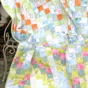 May include: A patchwork quilt with a variety of patterned squares in shades of green, orange, pink, and blue. The quilt is draped over a white metal chair, showcasing its intricate design and color palette. The quilt's edges are finished with a green and white striped border.