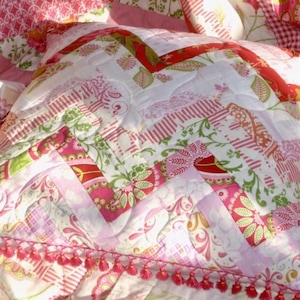 Op de afbeelding: Een roze en witte patchworkdeken met een bloemenpatroon. De deken heeft een gerimpelde rand en is gemaakt van verschillende stoffen.