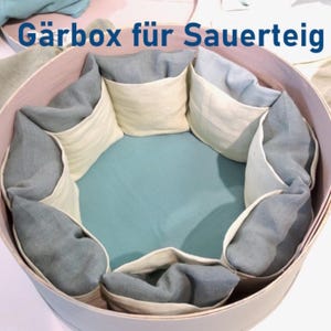 Peut inclure: Une boîte ronde en bois clair avec une doublure bleu clair. La boîte est divisée en sections avec des séparateurs en tissu. La boîte porte l'inscription "Gärbox für Sauerteig" sur le dessus.