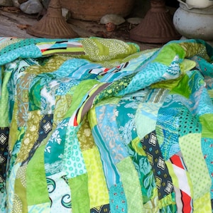 Può includere: Un quilt patchwork realizzato con diversi tessuti verdi, blu e bianchi. Il quilt è composto da molti motivi e texture diverse, ed è piegato in modo da mostrare i diversi colori e design.