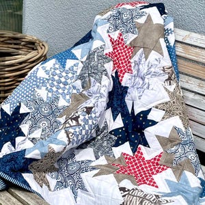 Könnte beinhalten: Eine handgefertigte Steppdecke mit einem Sternmuster in Blau-, Rot-, Braun- und Weißtönen. Die Steppdecke besteht aus verschiedenen gemusterten Stoffen, darunter Punkte und geometrische Designs. Die Steppdecke ist über einer Holzbank drapiert.