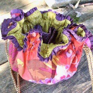 Può includere: Una borsa a cordoncino con un bordo a balze viola e verde. La borsa è realizzata in un tessuto floreale colorato con sfumature di rosso, arancione, giallo e rosa.