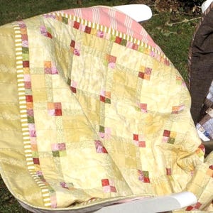 Könnte beinhalten: Eine gelbe, rosafarbene und grüne Patchwork-Decke mit einem weißen und rosafarbenen gestreiften Rand. Die Decke ist gefaltet und über einen Stuhl gelegt.