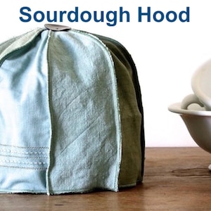 Könnte beinhalten: Ein hellblauer Stoffkorb zum Gehenlassen von Sauerteigbrot mit einem Metallgriff. Der Korb steht auf einer Holzoberfläche.