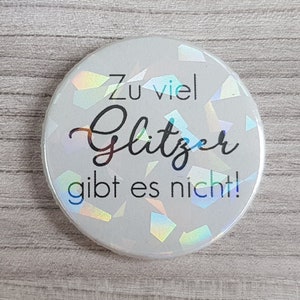 Könnte beinhalten: Ein runder, weißer Button mit schillernden Glitzerakzenten. Der Button zeigt den deutschen Text "Zu viel Glitzer gibt es nicht!" in schwarzer Schrift, was bedeutet "Es gibt nicht zu viel Glitzer!"