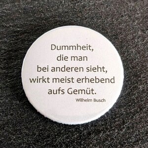 Könnte beinhalten: Ein weißer, runder Button mit schwarzem Text. Der Text lautet: "Dummheit, die man bei anderen sieht, wirkt meist erhebend aufs Gemüt. Wilhelm Busch."