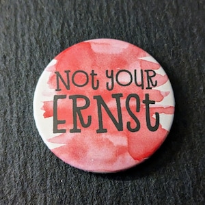 Könnte beinhalten: Ein runder Button mit einem Aquarelldesign in Rot- und Weißtönen. Der Button zeigt den Schriftzug "Not your Ernst" in schwarzen, fetten Buchstaben. Ein lustiges Accessoire.
