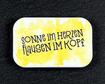 May include: A rectangular pin with rounded corners featuring a yellow watercolor background and black text that reads "SONNE IM HERZEN FLAUSEN IM KOPF" in a stylized font. The pin has a white border surrounding the yellow watercolor design.