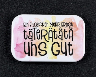 May include: A rectangular pin with rounded corners featuring a white background with soft pink and yellow watercolor splashes. The black text reads: Ein Bisschen Mehr Ernst, täterätätä, uns Gut. The design is centered on the white surface of the pin.