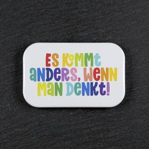Könnte beinhalten: Weiße rechteckige Dose mit einem regenbogenfarbenen Spruch, der "Es kommt anders, wenn man denkt!" lautet.