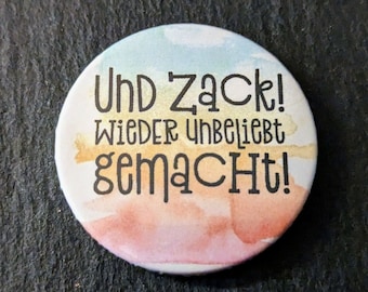 May include: A round button with a watercolor background in shades of blue, peach, and gold. The button features the German text "Und Zack! Wieder Unbeliebt gemacht!" in black, stylized lettering.