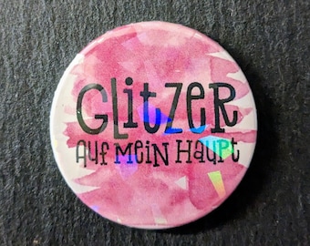 May include: A round button with the German text "GLITZER Auf Mein Haupt" in black lettering. The button has a watercolor-style background in shades of pink and white, with iridescent accents.