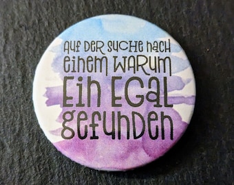 May include: A round button with a watercolor design in shades of blue and purple. The button features black text that reads "AUF DER SUCHE NACH EINEM WARUM EIN EGAL GEFUNDEN."