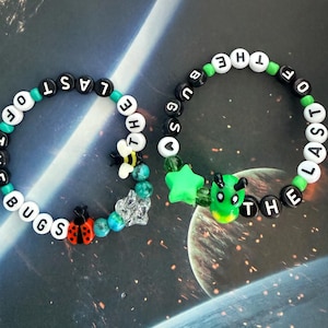 Puede incluir: Dos pulseras de cuentas con el texto "THE LAST OF THE BUGS". Una pulsera presenta un dije de mariquita, la otra un dije verde con forma de alienígena. Las cuentas son negras y blancas con detalles verdes y turquesas.