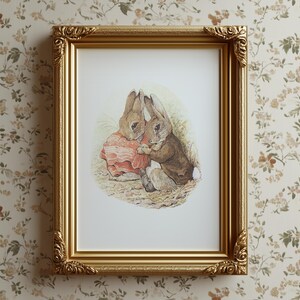 May include: A framed print featuring two rabbits in a classic illustration style. The rabbits are depicted in warm tones, with one wearing a brown jacket and the other a pink dress. The gold frame adds a touch of elegance.