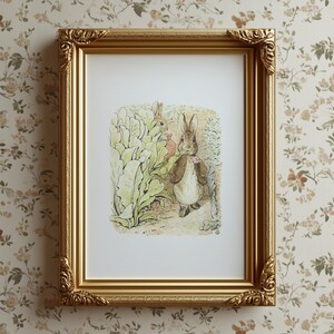 Puede incluir: Una impresión enmarcada con una ilustración clásica de un conejo con abrigo marrón y pajarita rosa, cerca de un jardín. El marco dorado tiene detalles ornamentados. La impresión se muestra sobre un papel pintado floral.