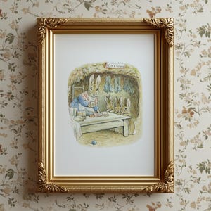 May include: A framed print featuring a watercolor illustration of rabbits in a cottage setting. The print shows a rabbit in a blue dress and apron, with three smaller rabbits. The gold frame has ornate detailing and is displayed against a floral wallpaper.