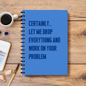 May include: A blue spiral-bound notebook with the text "CERTAINLY... LET ME DROP EVERYTHING AND WORK ON YOUR PROBLEM" sits on a wooden surface. A cup of coffee, art supplies, and a small notebook are also on the table.