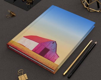 Könnte beinhalten: Ein Hardcover-Journal mit einer farbenfrohen Illustration eines Bauernhofs mit rotem Dach und blauem Himmel. Goldene Büroklammern, Bleistifte und ein dekoratives goldenes Objekt umgeben das Journal.