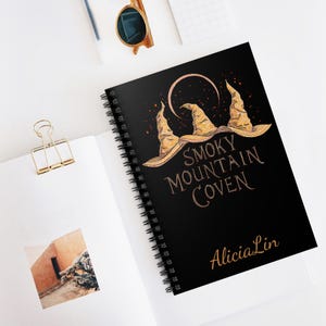 Pode incluir: Um caderno preto com espiral com as palavras "Smoky Mountain Coven" e "Alicia Lin" em letras douradas. A capa apresenta três chapéus de bruxa dourados e um design de lua crescente.