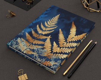 May include: A hardcover journal with a blue cover featuring a gold fern leaf design. The journal is open, revealing blank pages. Gold binder clips and pencils are arranged around the journal, creating a stationery theme.