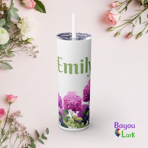 Op de afbeelding: Een witte beker met een rietje, met de naam "Emily" in groene letters. De onderkant van de beker is versierd met een bloemenontwerp van paarse hortensia's. Het Bayou Lark-logo is rechtsonder zichtbaar.