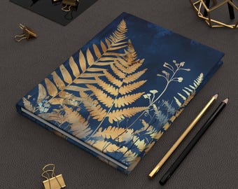 May include: A hardcover journal with a dark blue cover featuring a gold fern and botanical design. Gold and black pencils and gold binder clips are arranged around the journal on a gray surface. The journal is a nature-inspired stationery item.