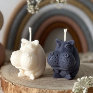 May include: Two small hippopotamus shaped candles, one white and one grey, sitting on a wooden slice. The candles are on a white surface with white flowers.