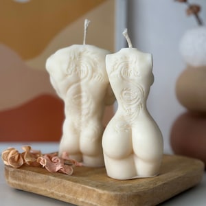 May include: Two white candles shaped like human torsos with a dragon design carved into the back. The candles are on a wooden tray.