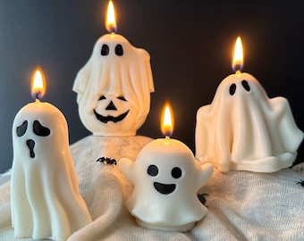 May include: Four white ghost-shaped candles with black facial features. The candles are lit and sitting on a white fabric surface.