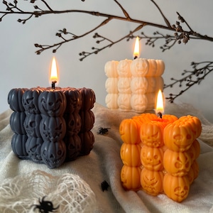 Può includere: Tre candele a tema Halloween, una nera, una bianca e una arancione, ciascuna a forma di cubo con più facce di zucca.