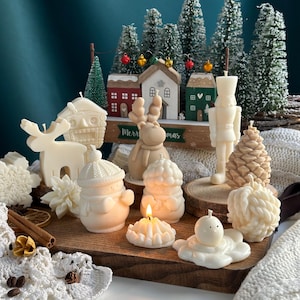 Puede incluir: Una colección de velas de temática navideña blancas y marrones, que incluyen un muñeco de nieve, un reno, una piña y una casa. Las velas están dispuestas sobre una superficie de madera con un mantel de ganchillo blanco y palitos de canela.