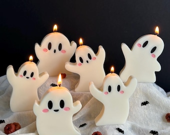 May include: Six white ghost-shaped candles with lit wicks. Each candle has a friendly face with black eyes and a pink blush. The candles are arranged on a white cloth with small decorative bats and pumpkins, perfect for Halloween.