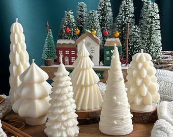 May include: A set of six white Christmas tree candles, each with a unique design. The candles are arranged on a wooden surface with a miniature village scene in the background.