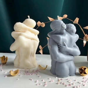 Op de afbeelding: Twee sculpturale kaarsen in de vorm van omhelzende figuren. De ene kaars is gebroken wit en de andere is leigrijs. Elke kaars toont twee personen in een nauwe omhelzing, met gedetailleerde ruggen, schouders en haartexturen. De lonten zitten boven.
