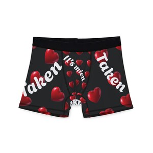 Peut inclure: Boxer short noir avec un motif répété de coeurs rouges. Les mots "Taken" et "It's mien" sont imprimés en blanc. La ceinture est noire.