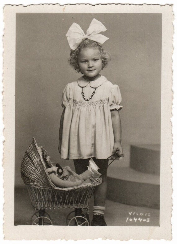 Little Girls Fashion Of The 1940s With Her Huge White Hair Ribbon Dress And Necklace. The Decor Is Completed With A Toy Pram And Doll Inside