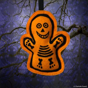May include: Orange felt Halloween decoration in the shape of a skeleton. The skeleton has black outlines and details, including a smiling face, rib cage, and bone-shaped hands and feet. A black ribbon is attached for hanging. The background features tree branches and a patterned design.