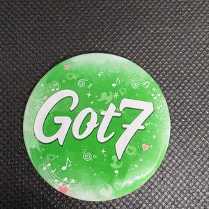 Könnte beinhalten: Ein runder, grüner Button mit dem weißen Text "Got7". Der Button zeigt Noten, Herzen und Sterne. Der Hintergrund hat einen Farbverlauf, der von einem dunkleren Grün zu einem helleren Farbton übergeht. Ein Fanartikel.