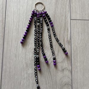 Könnte beinhalten: Ein Schlüsselanhänger mit mehreren Strängen aus schwarzen und lila Perlen. Die Stränge sind mit Buchstabenperlen verziert, die Sätze wie "PART OF ME" und "WHAT THE HELL YOU WANT FROM M" bilden. Der Schlüsselanhänger hat einen silbernen Ring.