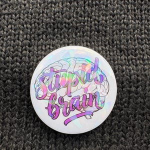 Könnte beinhalten: Ein weißer Button mit holografischer, irisierender Oberfläche. Er zeigt eine Gehirngrafik mit den Worten "Stupid Brain" in einer bunten, kursiven Schrift. Der Hintergrund ist ein dunkelgraues, strukturiertes Gewebe.