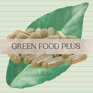May include: A collection of light brown capsules, labeled "GREEN FOOD PLUS", are arranged on a large green leaf. The capsules are transparent, revealing a green powder inside. The background is a light green color with subtle horizontal lines.